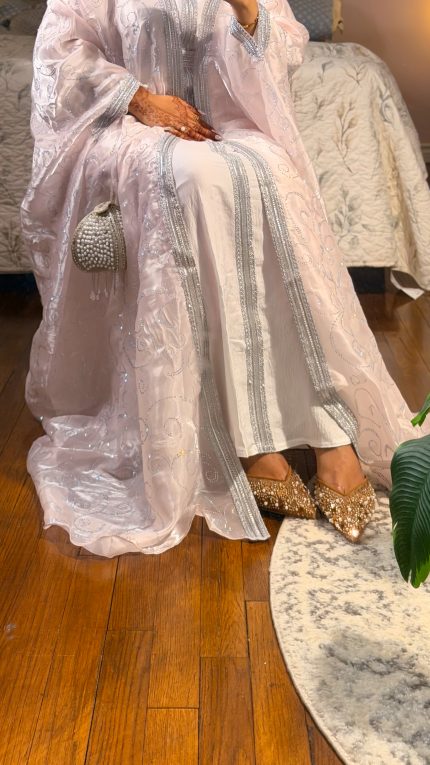 Woman wearing a subtle pink luxury organza abaya with all-over shimmering embroidery, styled with a white inner dress and matching hijab, elegant evening wear with soft flow and detailed finish.