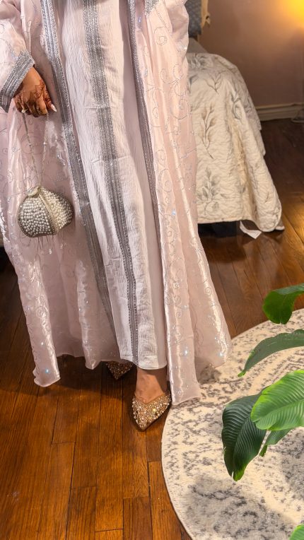 Woman wearing a subtle pink luxury organza abaya with all-over shimmering embroidery, styled with a white inner dress and matching hijab, elegant evening wear with soft flow and detailed finish.